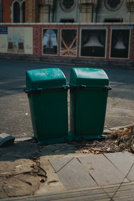 A middle-aged man with dark hair is seen collecting rubbish from a stainless steel trash bin on a paved sidewalk. He is wearing a black T-shirt with white and blue text and graphics, and dark trousers. The man is holding a large white plastic bag filled with waste in his left hand, while his right hand reaches into the open top of the bin to dispose of more rubbish. A wooden stick or broom handle is tucked under his arm, possibly used for assistance in rubbish collection. The scene is outdoors, with a background of lush green trees and a stone balustrade, indicating a public park or promenade. The lighting suggests daylight, with natural sunlight illuminating the area. This image relates to private and community waste collection, exemplifying independent rubbish clearance activities often undertaken to support local rubbish removal services like those offered by companies such as rubbishclearancebelsizepark.co.uk, emphasizing effective on-site rubbish disposal methods without reliance on municipal waste services.
