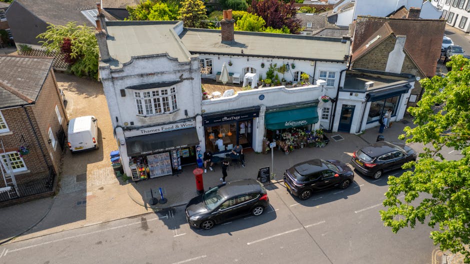 An aerial view of a small commercial street featuring three adjacent shops with white and light-colored exteriors. The shop on the left displays a sign reading 'Thames Food & Wine' and has a stand with newspapers and magazines positioned outside its entrance, alongside a traditional red post box nearby. Several pedestrians are walking or standing in front of the shops, some near parked cars on the street. In front of the shops, three black cars are parked along the curb, with one car slightly overlapping a designated parking space. To the left, a white van is parked on a side area paved with beige bricks, adjacent to old brick residential buildings. The buildings are surrounded by green trees, with sunlight casting shadows on the street and facades, indicating a clear day. The scene captures a typical village or small-town setting, potentially aligned with private waste collection and rubbish removal services working in the area, consistent with a guide to local rubbish clearance options in Belsize Village.