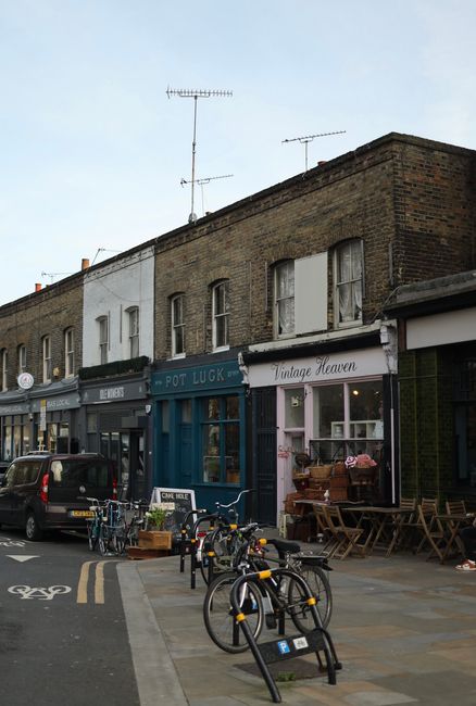 A row of small commercial buildings on a street features various shopfronts including a vintage store named 'Vintage Heaven' with a white sign and large display window, and a shop with a dark green facade and signage reading 'Rot LUCK'. Several bicycles are secured to black bike racks on the pavement in the foreground. To the left, a black car is parked, partially blocking a designated cycling lane marked with a white bicycle symbol and double yellow lines along the curb. The street appears active with outdoor seating visible outside the vintage shop, consisting of wooden tables and chairs. The upper floors of the brick buildings have small rectangular windows, some with open shutters or curtains, and the building facades show a mix of brickwork with a weathered appearance and painted sections. Atop the buildings, television antennas extend upwards against a pale blue sky with sparse cloud cover. The scene suggests a typical urban area with mixed-use shops, supporting local businesses and pedestrian activity, reflective of alternative waste management options, as exemplified by the shops occupying the premises. The overall atmosphere is calm, with natural daylight illuminating the scene, capturing everyday street life and commercial tenancy.