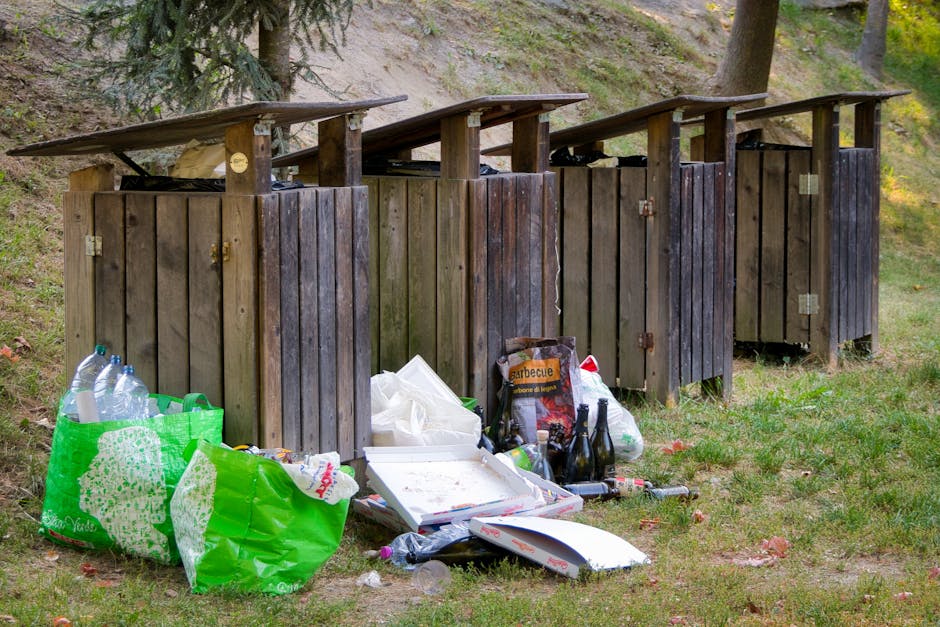 A row of four wooden outdoor rubbish bins with slanted roofs, positioned on a grassy area near a hillside with trees, showing signs of weathering with some darkened and aged wood panels. In front of the bins, there are multiple bags of waste, including a large green plastic bag filled with empty plastic bottles, a cardboard box containing discarded food containers, and smaller transparent and opaque plastic bags. Several glass bottles and a few scattered beverage cans are lying on the ground beside the waste collection, along with a plastic wrapper and other small debris. The scene suggests a typical outdoor waste disposal site, possibly used for household rubbish, with natural surroundings and minimal human activity, and could be associated with private rubbish clearance services such as those offered by rubbishclearancebelsizepark.co.uk.