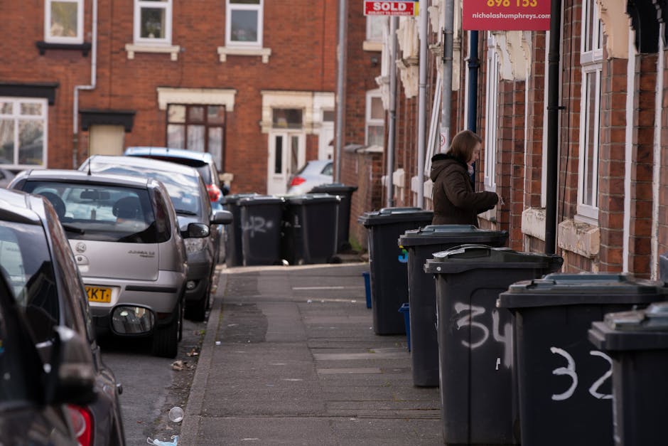 A black, cast-iron outdoor rubbish bin situated on a paved urban street with cobblestones, surrounded by a substantial amount of litter. The bin's lid is partially open, and various types of waste are placed on top, including paper cups with plastic lids, a plastic container, and miscellaneous food packaging. The base of the bin is cluttered with empty glass bottles, some still capped, and additional scattered trash, such as crumpled paper and disposable gloves, nearby on the ground. In the background, there are blurred figures of pedestrians, parked cars, and other urban elements, indicating a busy street scene. The environment appears to be in a city or town center, with natural lighting suggesting daytime. The scene exemplifies a typical instance of urban littering, which can be addressed through private waste removal services like those offered by rubbishclearancebelsizepark.co.uk, highlighting the importance of proper rubbish handling and disposal for maintaining cleanliness in built-up areas.