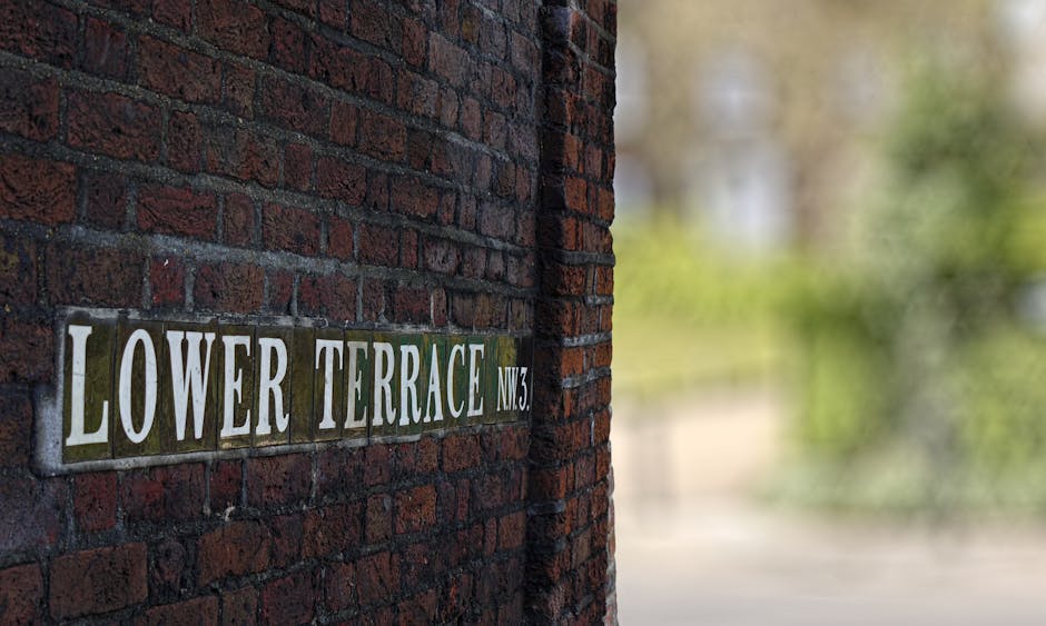 A close-up view of a street sign mounted on a red brick wall, reading 'Lower Terrace NW3' with the lettering in white against a dark green background. The sign consists of individual rectangular ceramic tiles, each bearing part of the text, arranged horizontally along the textured brick surface. The bricks exhibit a rough, weathered finish with visible mortar joints between them. Behind the wall, the background is blurred, revealing hints of greenery and soft natural light, suggesting an outdoor setting in a residential area. The lighting highlights the uneven texture of the bricks and the glossy surface of the tiles, emphasizing the sign's detail. The image is indicative of a quiet, well-maintained street, where private waste handling or on-site rubbish clearance might be typically carried out, aligning with the themes of independent rubbish removal services.